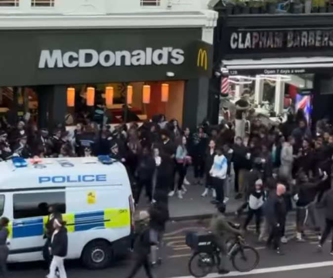 A crowd gathered in front of a McDonald's and Clapham Barbers, with a police van nearby and a person on a bicycle in the foreground.