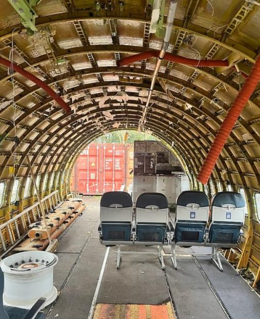 Interior of a stripped Boeing 737 fuselage with four seats installed, showing the exposed framework and red cargo containers outside.