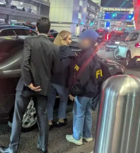 Woman with blonde hair standing near a black car, with a man in a suit and another person in a jacket with "FBI" on it.