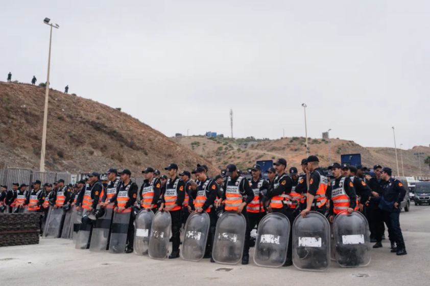 Police officers in black uniforms with orange vests stand in rows, holding riot shields.