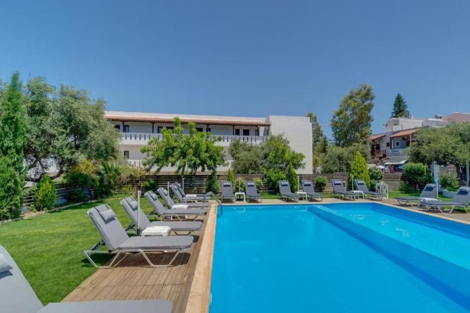 A bright blue swimming pool with rows of gray lounge chairs on a wooden deck and grass, with a white building and green trees in the background under a clear sky.