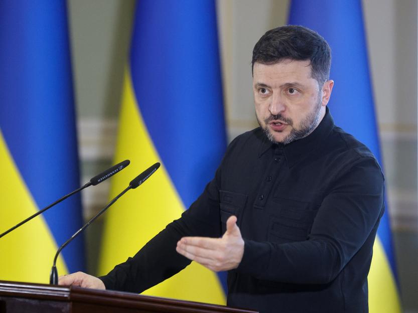 Ukrainian President Volodymyr Zelensky addressing a press conference in Kyiv, with Ukrainian flags in the background.