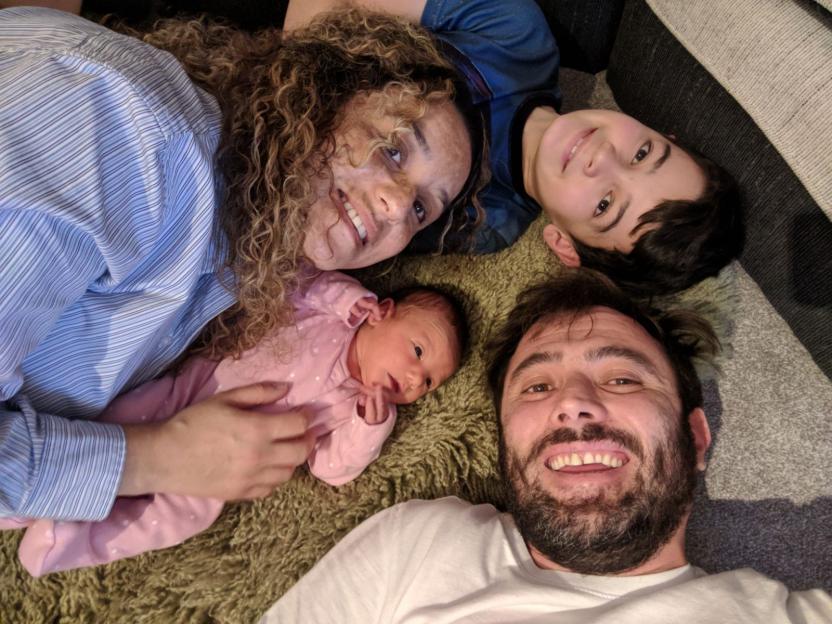 A family of four, a mother, father, boy, and newborn in a pink onesie, laying together on a carpet, smiling at the camera.