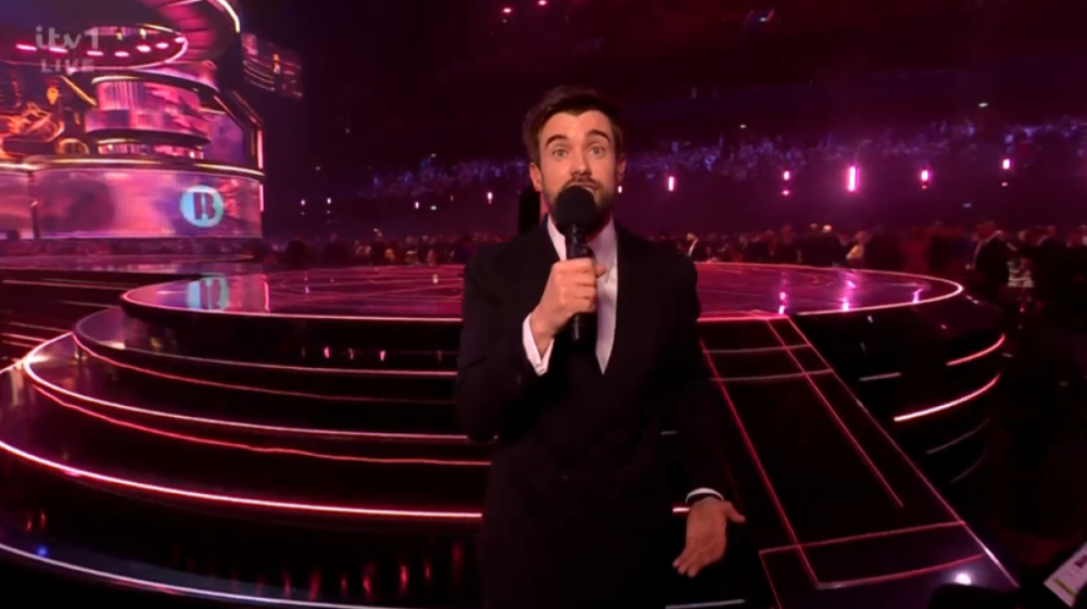A male host in a black suit with a microphone standing on a stage with glowing pink neon lights.