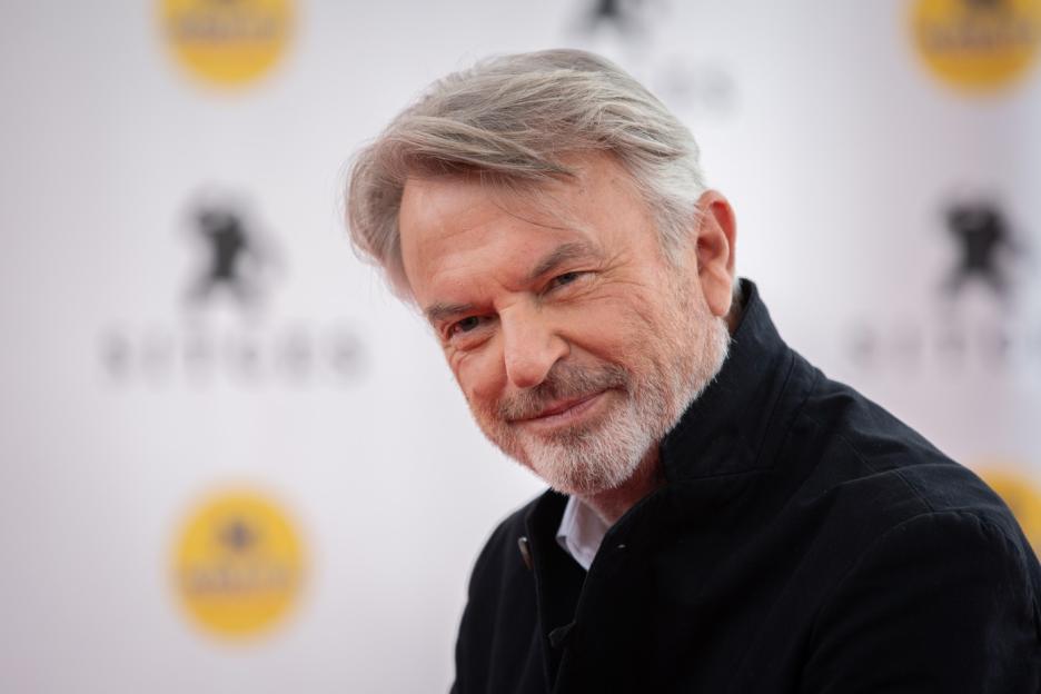 Actor Sam Neill with a gray beard and hair, wearing a black collared shirt, smiles and looks at the camera.