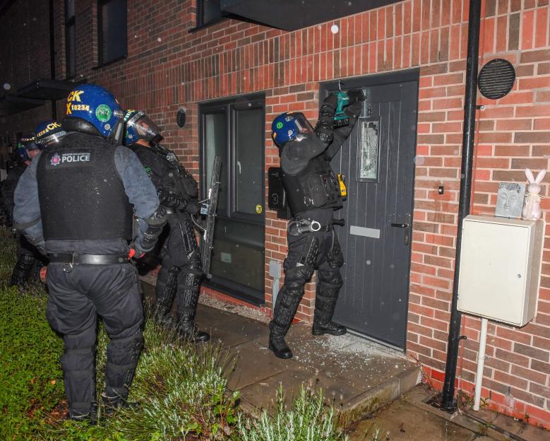 Police officers in tactical gear using a power tool to force entry into a home.