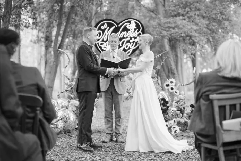 Black and white photo of Rufus Hound and his bride getting married outdoors, officiated by a man reading from a book.
