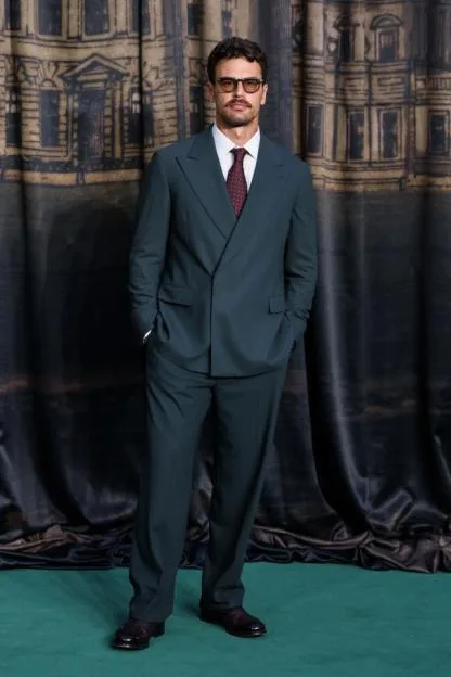 Theo James wearing a dark green suit, striped shirt, and patterned tie with glasses and a mustache.