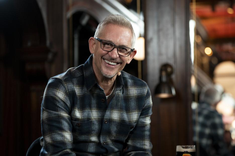 Gary Lineker, a white man with gray hair and glasses, smiles while wearing a plaid shirt.
