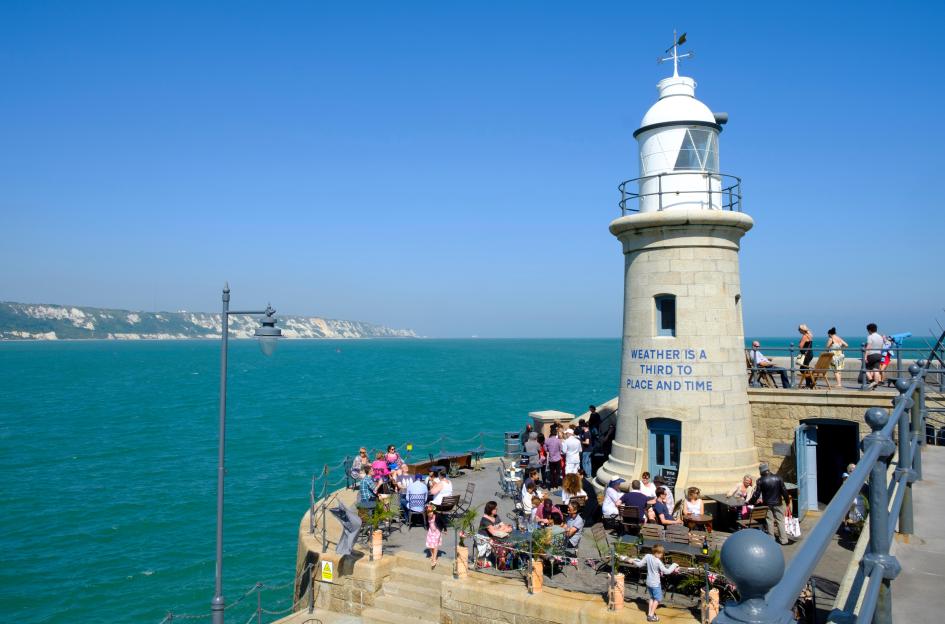 People enjoying drinks at an outdoor cafe next to a lighthouse on a sunny day.