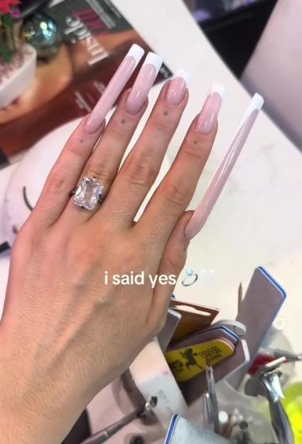 A woman's hand with extremely long square-tipped nude French manicured nails wearing a large rectangular engagement ring with the text "i said yes" above a small ring emoji.