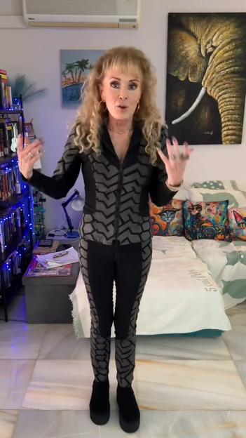 Beverley Callard wearing a dark gray and black patterned tracksuit, standing in a room with a bed and a bookshelf.