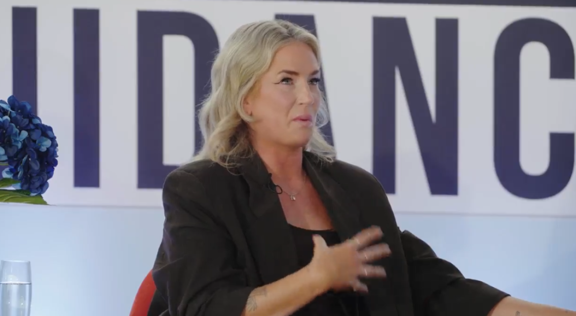 A blonde woman in a black jacket speaking during an event with a "Guidance" banner in the background.
