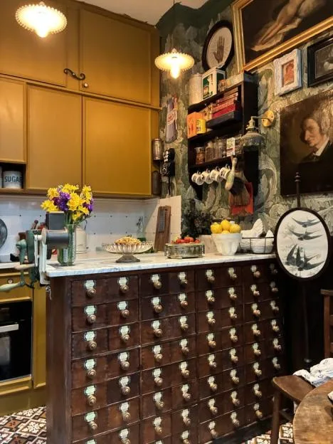 A kitchen with a vintage apothecary cabinet as an island, featuring mustard-yellow upper cabinets, floral wallpaper, and framed art.