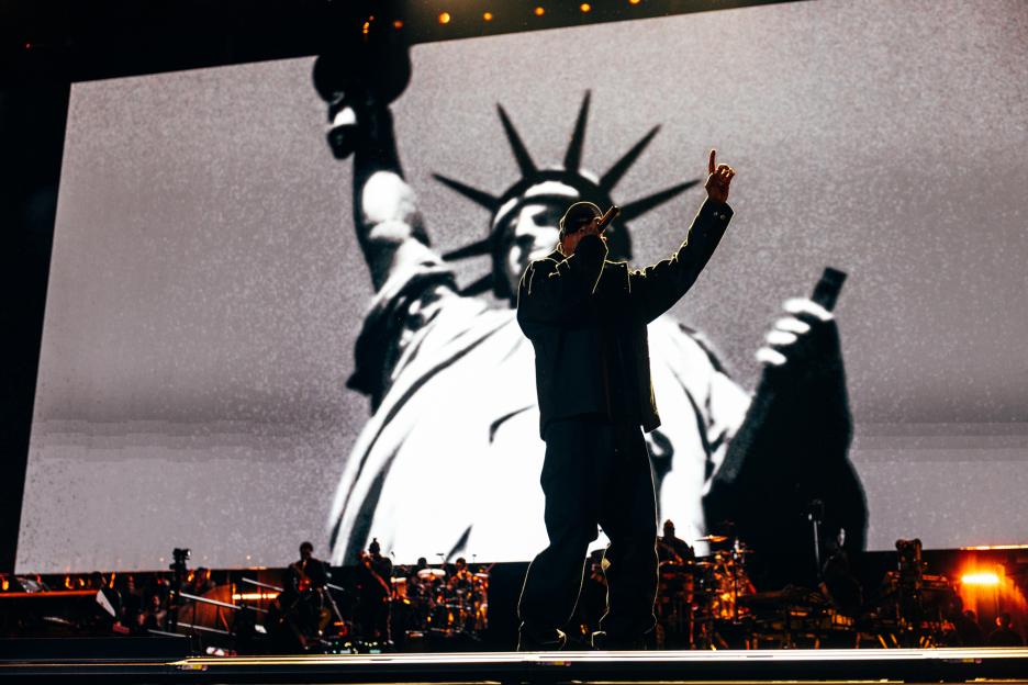 Jay-Z performing on stage with a large black and white image of the Statue of Liberty behind him.
