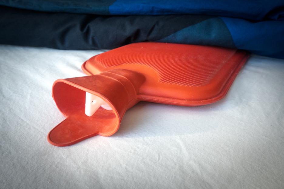 A red hot water bottle partially covered by a dark blue blanket on a white bed sheet.