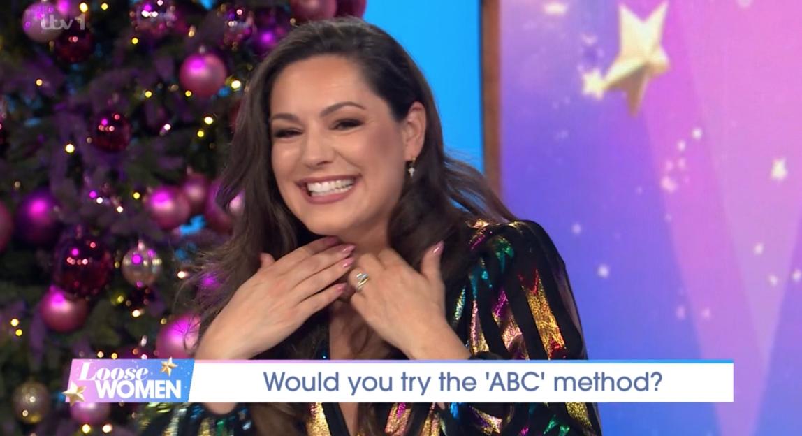 Kelly Brook smiling and holding her throat while wearing a sparkly outfit, in front of a Christmas tree.