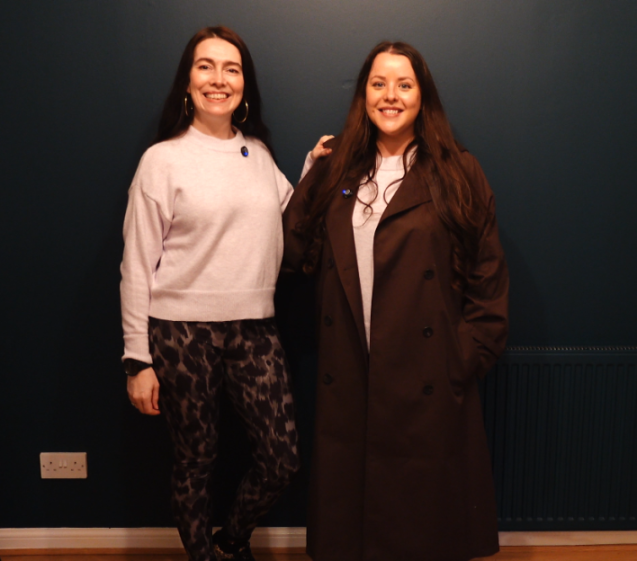 Two women wearing blue pins, standing and smiling at the camera.