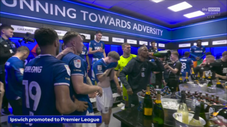 Ipswich Town Football Club players celebrate their promotion to the Premier League in a locker room, with one player drinking directly from a champagne bottle.