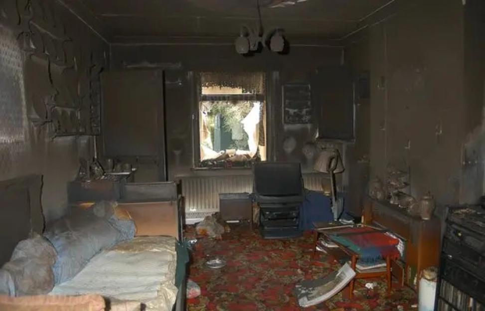 A bedroom with fire damage, walls and ceilings are blackened with soot, a broken window, and debris on the floor.