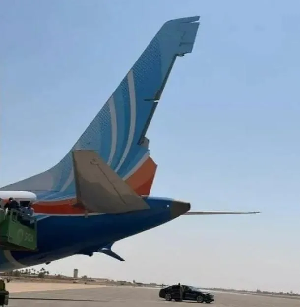 Rear section of a parked airplane on a tarmac with a car and people in the background.