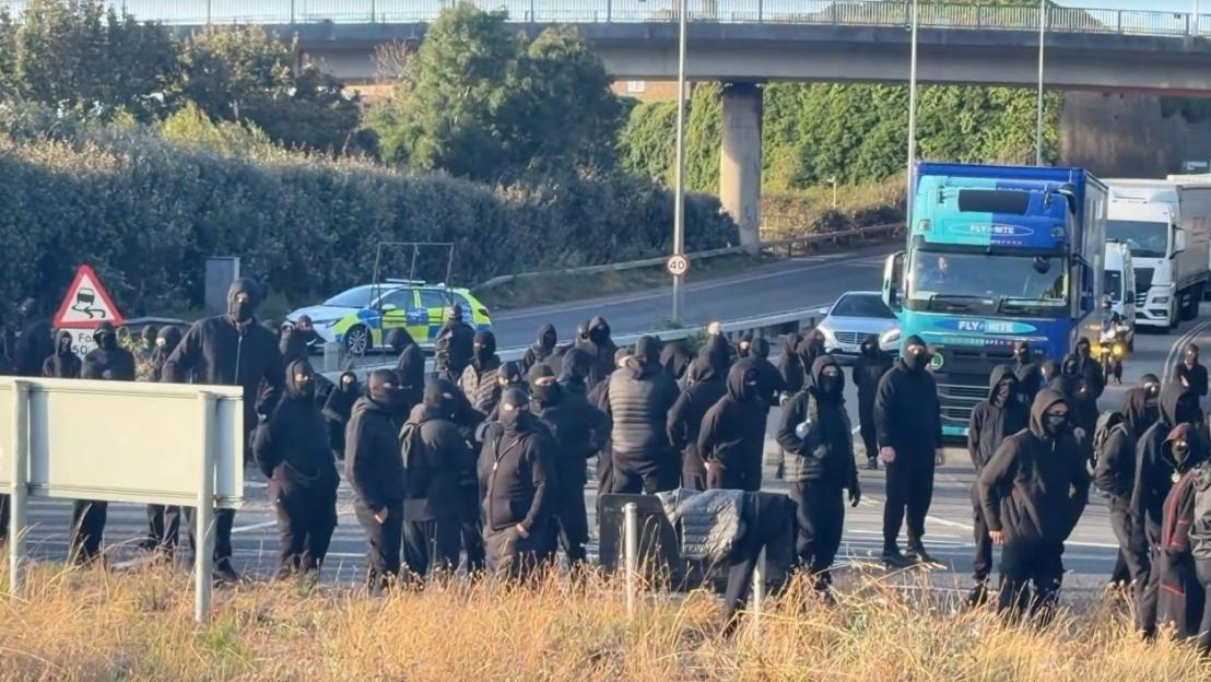 A group of protestors in black clothing and balaclavas standing on the side of a road in Dover, with a police car and trucks in the background.
