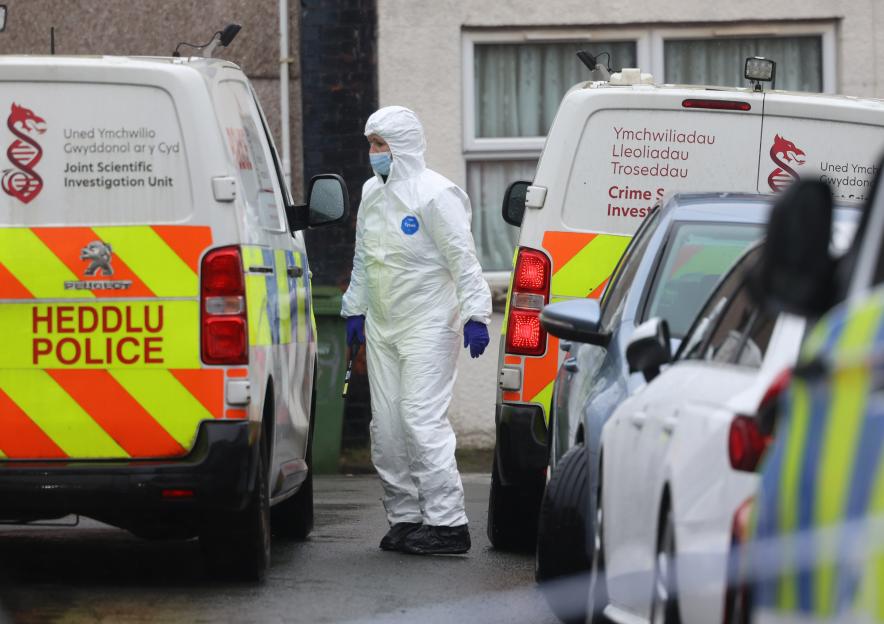 A Gwent Police officer in a white forensic suit and blue gloves stands between two police vans at a crime scene.