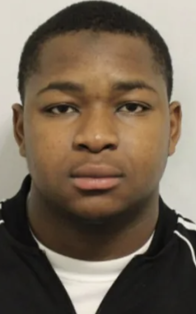 A man with dark skin and short dark hair looks directly at the camera. He wears a dark track jacket over a white t-shirt.