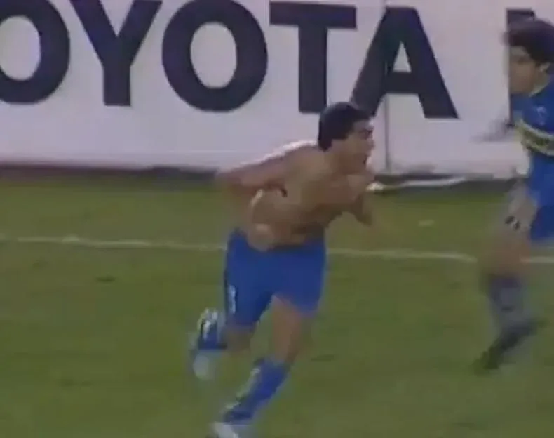 A shirtless soccer player in blue shorts running on the field, celebrating with another player in the background.