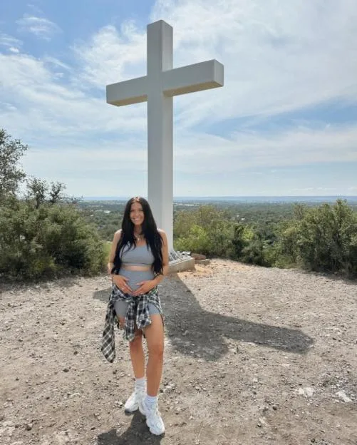 A pregnant woman standing outdoors in front of a large white cross.