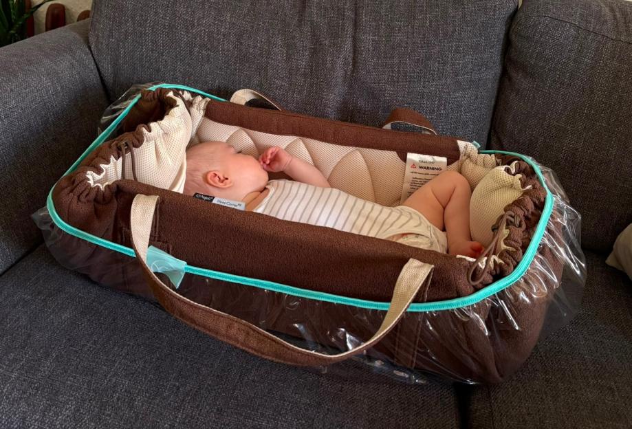 A baby sleeps soundly in a brown and turquoise SleepCarrier portable bassinet on a gray couch.