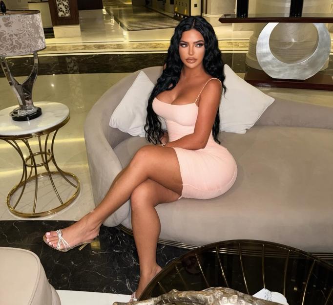 Woman with dark wavy hair in a light pink dress seated on a sofa.