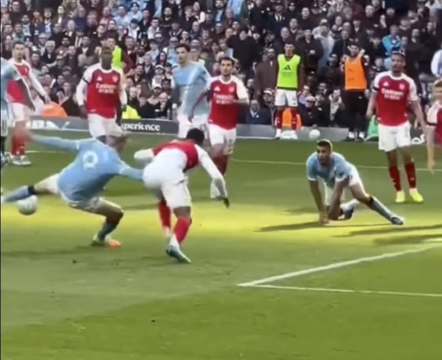 A soccer player in a light blue uniform falls backward as he attempts to kick the ball away from a player in a red uniform.