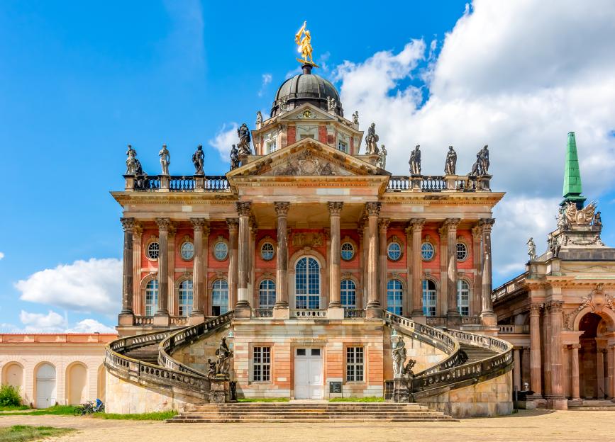 The New Palace in Potsdam, Germany, a red Baroque-style building with a domed roof topped by a golden statue and ornate sculptures along the roofline, is featured under a blue sky with white clouds.