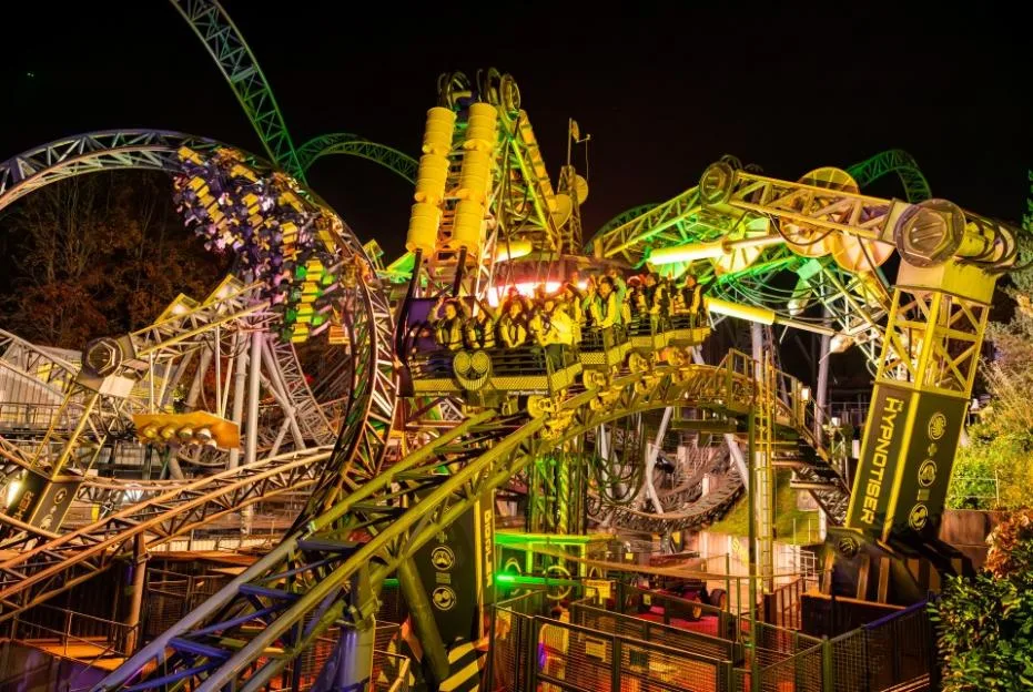 Rollercoaster cars filled with screaming riders navigate a complex network of illuminated steel tracks at night.