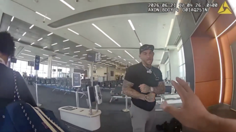 A man with tattoos on his arms, wearing a black shirt and baseball cap, appears to be speaking to someone in an airport terminal with empty seats.