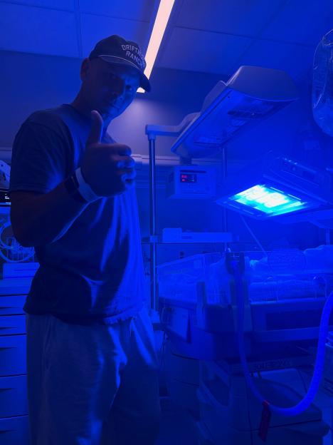 Man in a baseball cap giving a thumbs-up next to an incubator with a baby under blue lights.