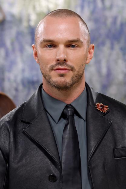 Nicholas Hoult in a dark shirt and tie with a black leather jacket and an ornate brooch on his lapel.