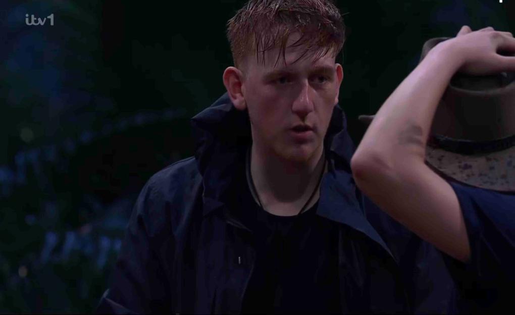 Angry Ginge in a dark hooded jacket looks at another person.