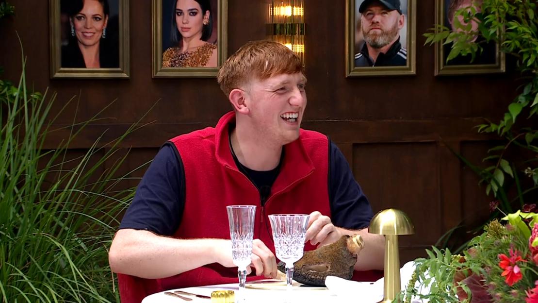 A man in a red vest and black shirt laughing at a dining table with crystal glasses, a golden mushroom lamp, and a taxidermy animal.