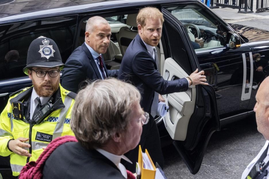 Prince Harry exiting a dark vehicle, escorted by a man in a suit and a police officer.