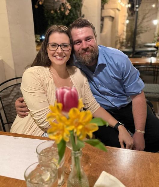 A smiling woman in glasses and a man with a beard sitting at a table with flowers in the foreground.