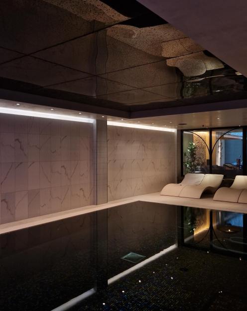 An indoor spa pool with two lounge chairs by the side, illuminated by warm lighting and reflections from the metallic ceiling.