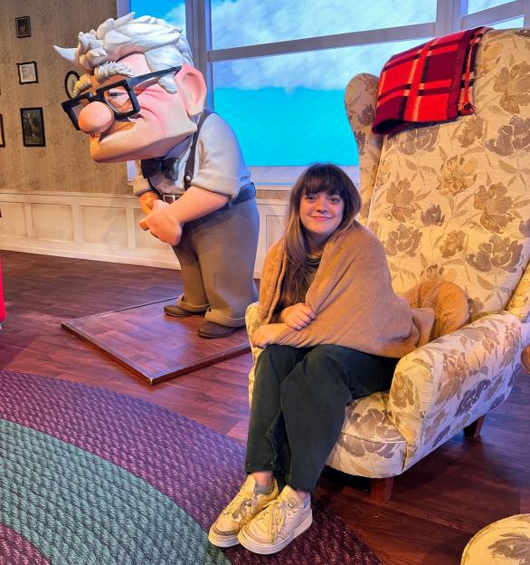 A person wrapped in a blanket sits in a floral armchair next to a life-sized figure of Carl Fredricksen from Pixar's Up.