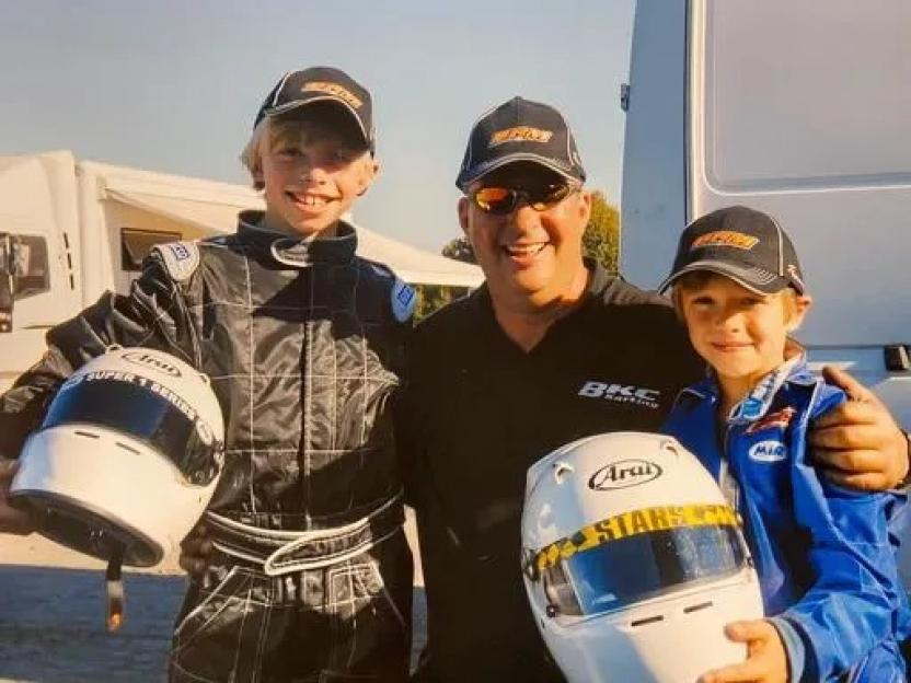 A man and two boys, all wearing racing gear and hats, smiling and holding helmets.