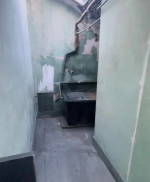 A narrow, run-down corridor with peeling green walls, exposed pipes, and a skylight at the end.