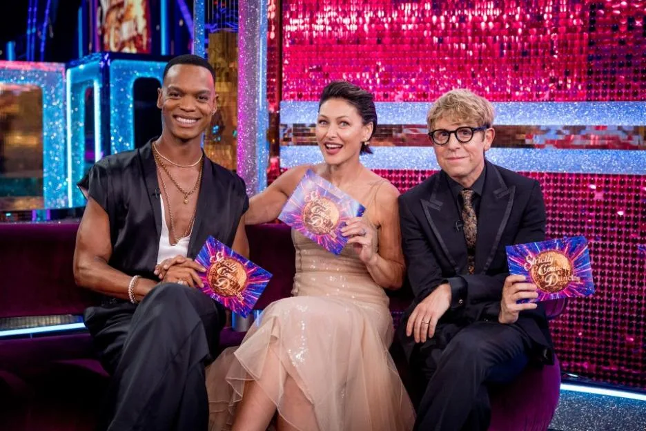 Strictly Come Dancing hosts Johannes Radebe, Emma Willis, and Josh Widdicombe pose together on a couch, each holding a show card.