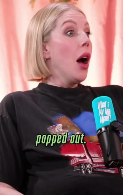 A woman with blonde hair, wearing a black t-shirt, talks into a microphone while her mouth is open in surprise.