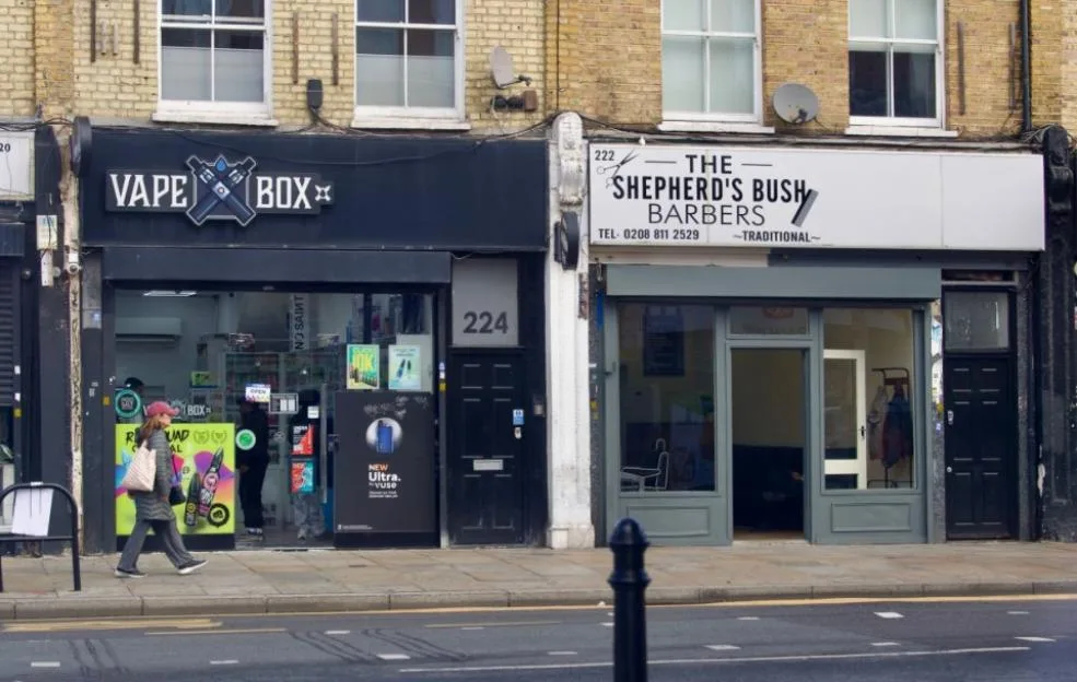 A vape shop and a traditional barbershop storefronts on Uxbridge Road in London.