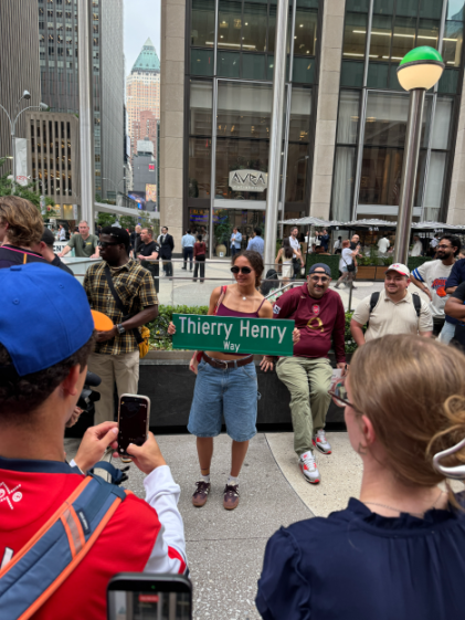 A crowd of people on a city street, with a woman in the center holding a "Thierry Henry Way" sign, as others take photos and look on.